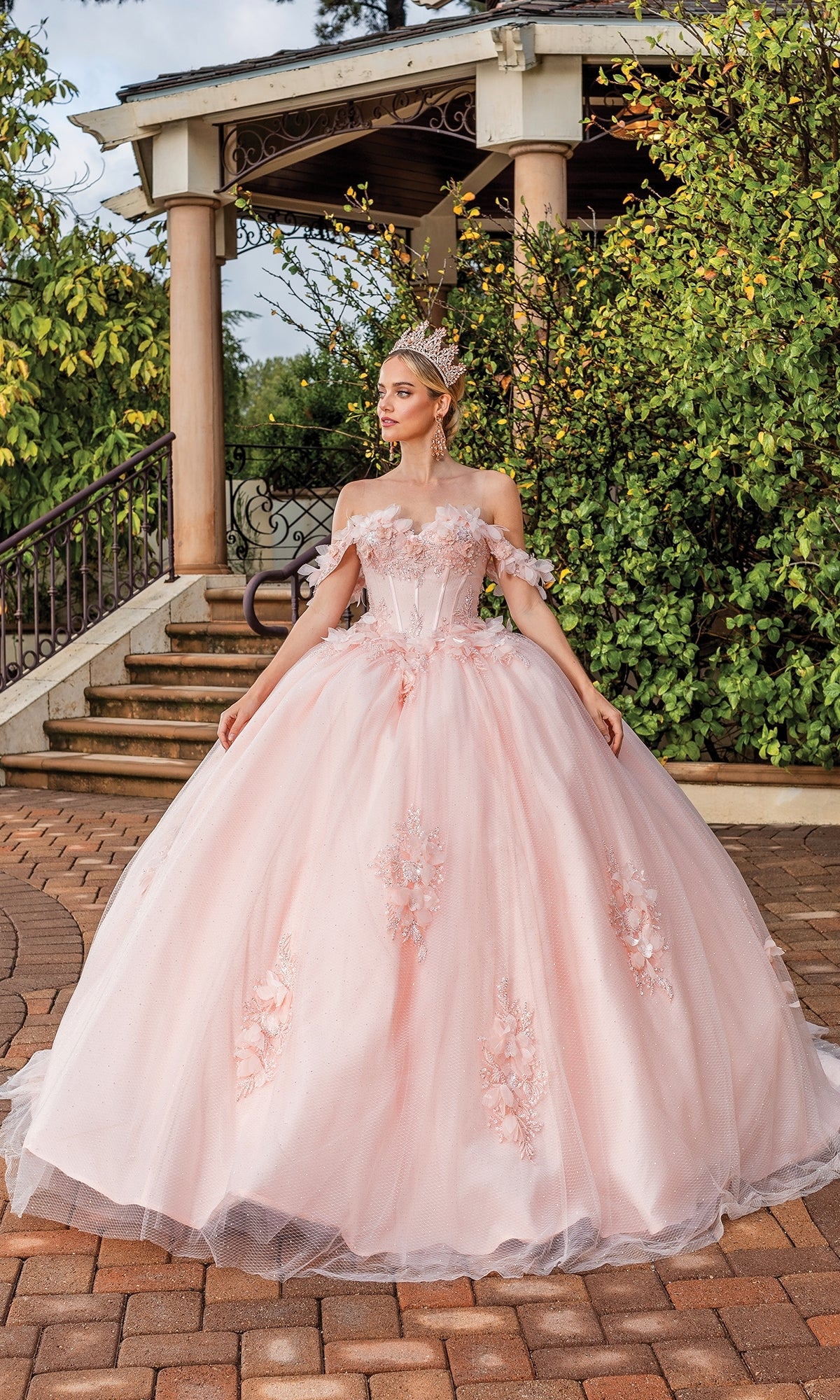 A woman in the Quinceanera Dress 1883 By Dancing Queen, a pink off-the-shoulder A-line ball gown, with a crown, stands on a brick pathway before a gazebo surrounded by greenery.